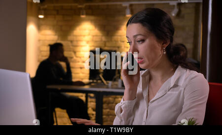 Closeup shoot of young pretty caucasian businesswoman having a phone call while sitting in front of the laptop indoors on the workplace Stock Photo