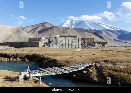 LAKE KARAKUL, XINJIANG / CHINA - October 2, 2017: Mount Muztagata with traditional houses, a river and a wooden bridge in the foreground. Stock Photo