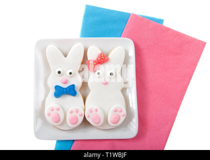 Easter bunny cookies, boy and girl with bow and bow tie on a small square plate sitting on pink and blue napkins isolated on white background. Close u Stock Photo