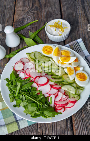 healthy low calories spring salad of wild garlic, hard boiled eggs, radish and cucumber on a white plate on a rustic wooden table with yogurt sauce in
