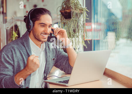 Man smiling having a online conversation looking at laptop screen, cheering, pumping fist gesticulating at home in living room, near window holding he Stock Photo