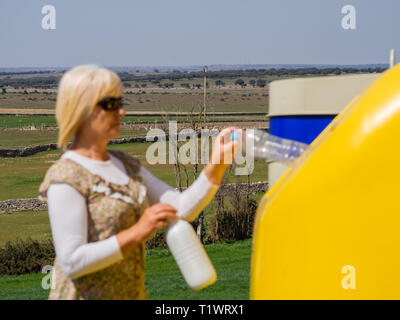 A mature woman pulling a plastic bottle in a yellow bin for recycling plastic Stock Photo