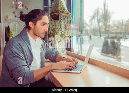 Man typing at laptop. Portrait of a handsome guy wearing formal white shirt and gray blouse sitting near window at table in living room or coffee shop Stock Photo