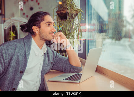 Man daydreaming in front of the computer. Closeup portrait of a handsome guy wearing formal clothing looking through window at a table in living room  Stock Photo