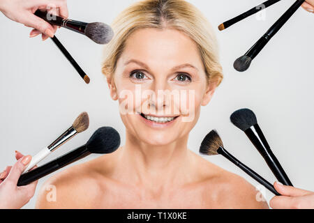beautiful and smiling woman looking at camera and makeup artists holding cosmetic brushes isolated on grey Stock Photo