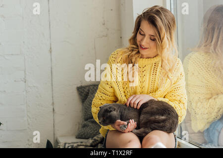 beautiful smiling young woman in knitted sweater hugging scottish fold cat while sitting on window sill Stock Photo