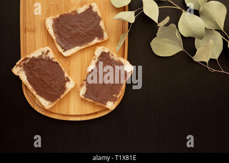 A composition of three white toasts smeared with chocolate butter that lie on a cutting board against with leaves a dark background. top view with are Stock Photo