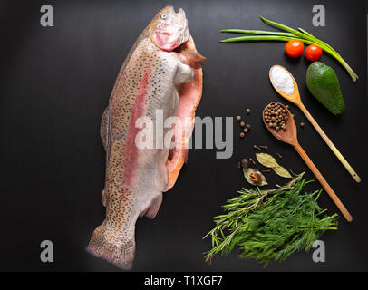 Whole raw filleted salmon trout with assorted herbs, seasoning, spices, tomato and spring onion on black viewed from above Stock Photo