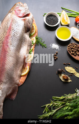 Fresh ingredients for preparing fresh salmon trout with aromatic herbs, spices, lemon, oil, and tomato viewed vertically high angle on a dark backgrou Stock Photo