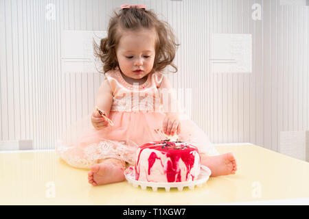 Little girl eyeing up her colorful birthday cake topped with red jelly as she sits holding part of the decoration with sticky fingers, dirty mouth and Stock Photo