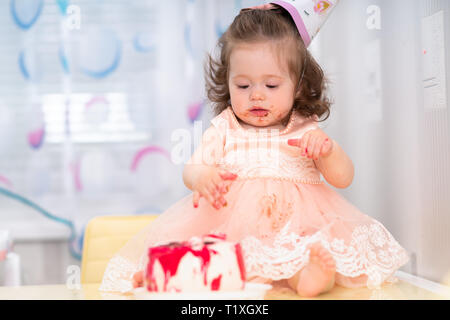 Cute sticky messy little girl in a pink dress eating her birthday cake wearing a party hat Stock Photo