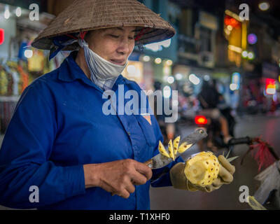 Vietnamese woman with classic conical hat preparing and slicing pineapple at night market in Hanoi, Vietnam Stock Photo