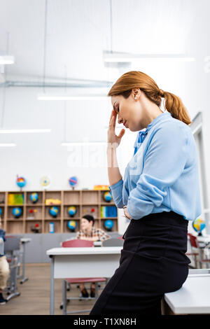 Tired teacher in blue blouse standing near table in classroom Stock Photo