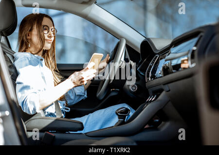 Young and cheerful woman using smart phone while sitting in the modern car in the city Stock Photo