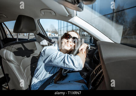 Young and cheerful woman enjoying new car hugging steering wheel sitting inside, wide angle view Stock Photo