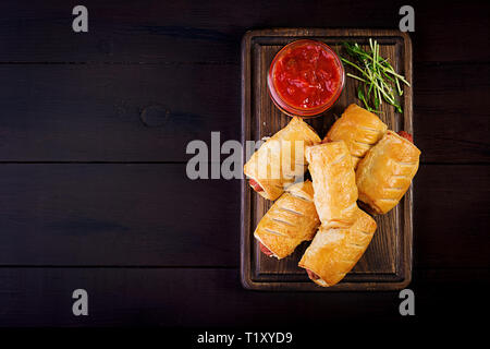 Sausage in the dough on a wooden board with tomato sauce. Top view Stock Photo
