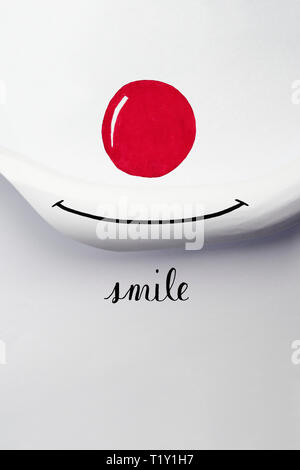 White paper is rolled up and forms a smiling mouth. A drawn red nose and a hand written word in english meaning: smile make a good mood. Stock Photo