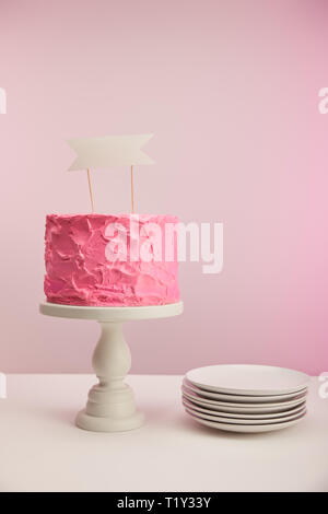 tasty and sweet birthday cake with blank card on cake stand near saucers on pink Stock Photo