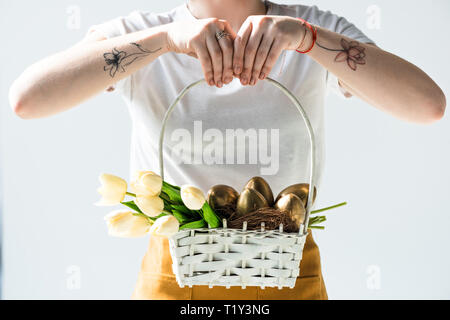 cropped view of woman holding straw basket with golden Easter eggs and tulips isolated on white Stock Photo