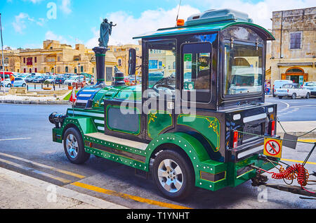 RABAT, MALTA - JUNE 16, 2018: The old-style fun train waits the tourists in Museum square of Rabat with monument of St Joseph and historical housing o Stock Photo