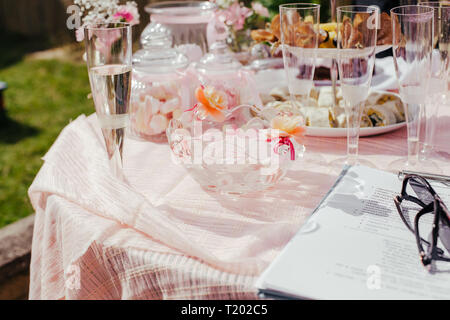 Candy table, sweets in the glass jars, pink decor on table outside in the summer. Party table with food, canapes, snacks. Planning party, notebook and Stock Photo