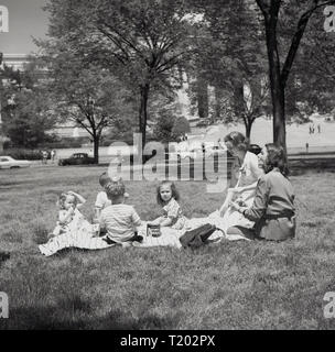 1950s, summertime and two mothers with their young children sitting outside on a rug having a picnic in the park infront of the Capitol building, Washington DC, USA, home to the US Government. Stock Photo