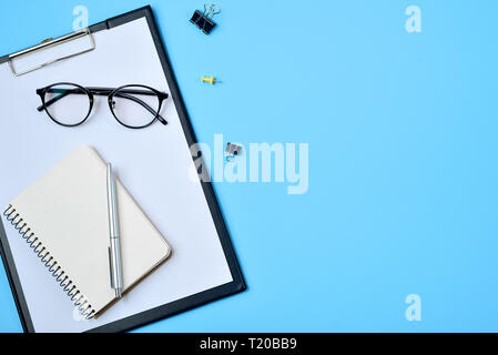 Flat lay, top view office table desk. Workspace with blank clip board, office supplies, pen, notepad, eyeglasses on blue background. Copy space, top v Stock Photo