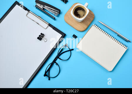 Office workspace with blank clip board, office supplies, pen, notepad, eyeglasses, stapler, coffee cup and spectacles on blue background. Flat lay, to Stock Photo