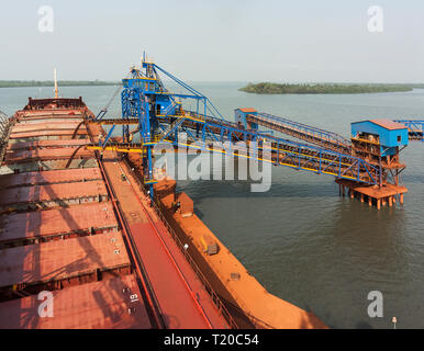 Port operations for managing and transporting iron ore. Loading holds of transhipper boat with ore via 2 ship loaders on jetty before transport to OGV Stock Photo