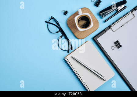 Office workspace with blank clip board, office supplies, pen, notepad, eyeglasses, stapler, coffee cup and spectacles on blue background. Flat lay, to Stock Photo