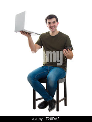 Full length portrait of casual young man sitting on chair holding laptop in one hand and mobile phone in another, comparing gadgets isolated over whit Stock Photo