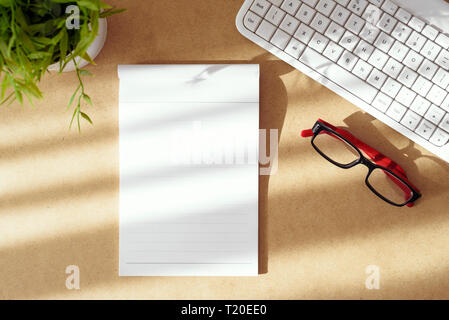 Top view of work place arrangement on desk table. Empty paper notepad, eye glasses, white computer keyboard and green plant. Copy space. Stock Photo