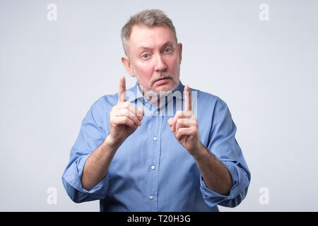 Experienced senior man in blue shirt showing index fingers up, giving advice or recommendation. Warning sign at studio shoot Stock Photo