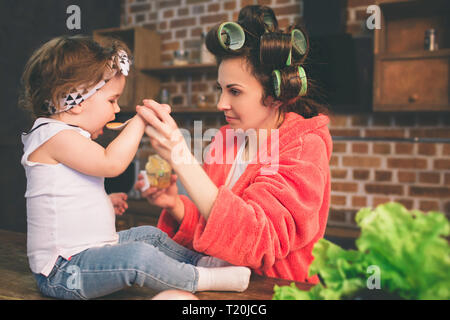 Mum at home. Young mother with little child in the home kitchen. Woman doing many tasks while looks after her baby. Stock Photo