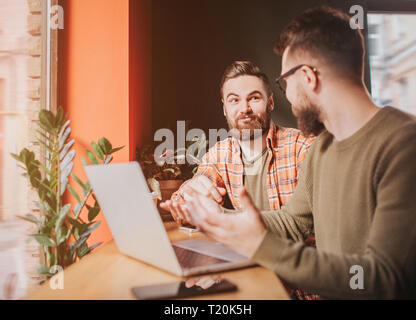 Close up of two bearded guy sitting at the table in front of the window. They are looking to each other and having conversation Stock Photo