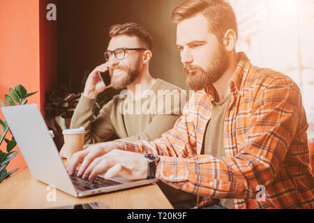 Busy and serious businessmen are woking in cafe. One of them is working on computer while another one is talking on the phone Stock Photo