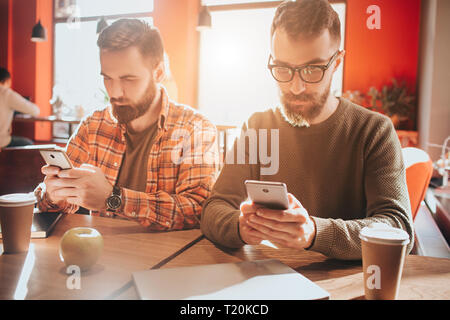 Take a look on beautiful and handsome bearded men sitting at te table and holding phones. They are looking only at their screens. Guys are busy. Close Stock Photo
