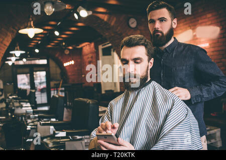 Bearded guy is sitting in chair and getting ready for procedure. He is looking at the phone. He is very concentrated. Hairdresser is ready to start pr Stock Photo