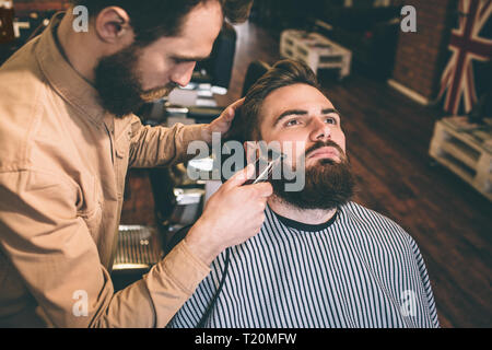 Nice and good hairstylist is cutting some of the customer's beard with an electrical razer. He is doing that very accurate Stock Photo