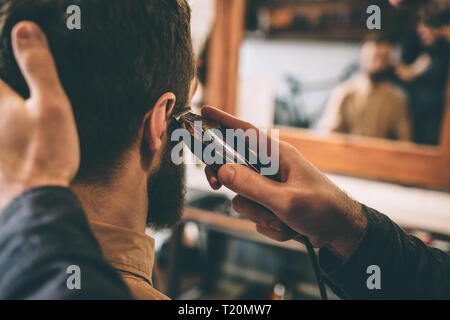 Cut view of guy cutting the hair of the customer with an electrical razer near right ear. He is holding a head and doing that very accurate Stock Photo