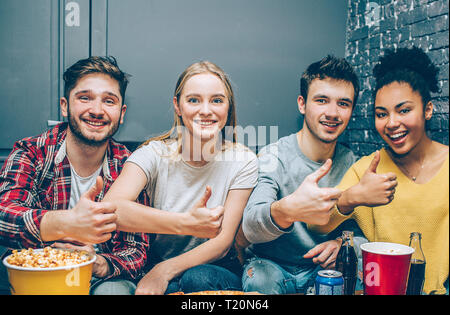 Close up of four best friends sitting together in a small room and posing to camera. They are showing their thumbs up and smiling. Teens look happy an Stock Photo
