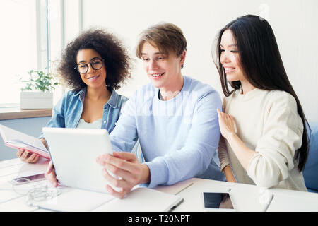Three beautiful students are sitting at the table and looking to the tablet's screen. They are studying together and preparing for exam Stock Photo