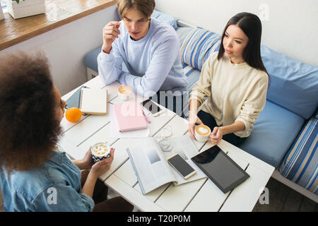 Nice picture of three students sitting at the table. There are lots of devices and notebooks on the table. Each of them has a cup of coffee. They are  Stock Photo