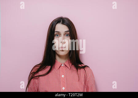 Studio portrait female keeps lips rounded, looks with positive expression, dressed in shirt. Beautiful student amazed to hear about coming session. Stock Photo