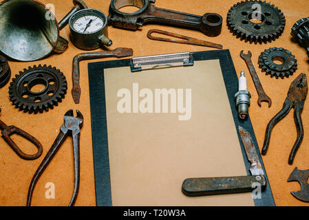 Clipboard with paper for your info in the center of rusty tools, gears on vintage fiberboard background. Motorcycle equipment and repair template. Stock Photo