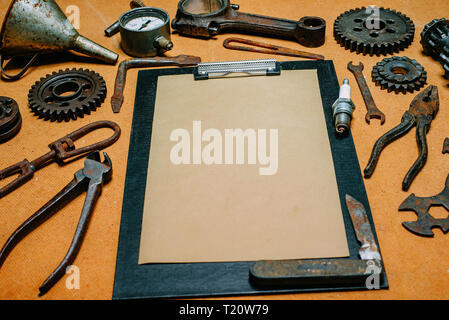 Closeup clipboard with paper for your info in the center of rusty tools, gears on vintage fiberboard background. Motorcycle equipment and repair templ Stock Photo