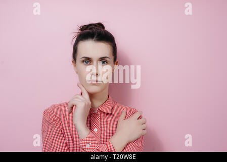 Horizontal portrait of lovely female student with hair knot, holds chin, wears shirt, looks seriously and with shy expression at camera, isolated on p Stock Photo