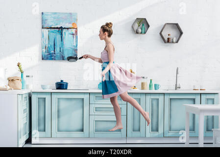 attractive barefoot young woman in apron levitating in kitchen while holding pan Stock Photo