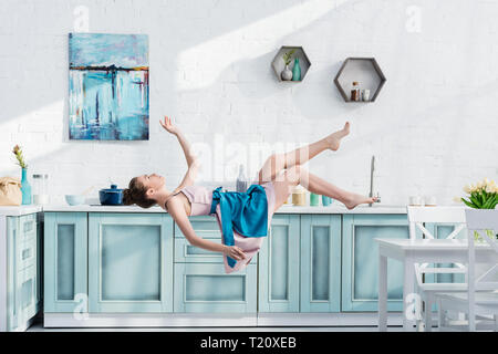 young elegant woman in apron and dress levitating in air in kitchen Stock Photo