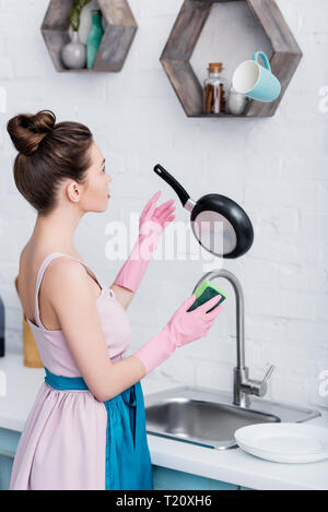 young woman in rubber gloves looking at levitating pan and cup in kitchen while washing dishes Stock Photo